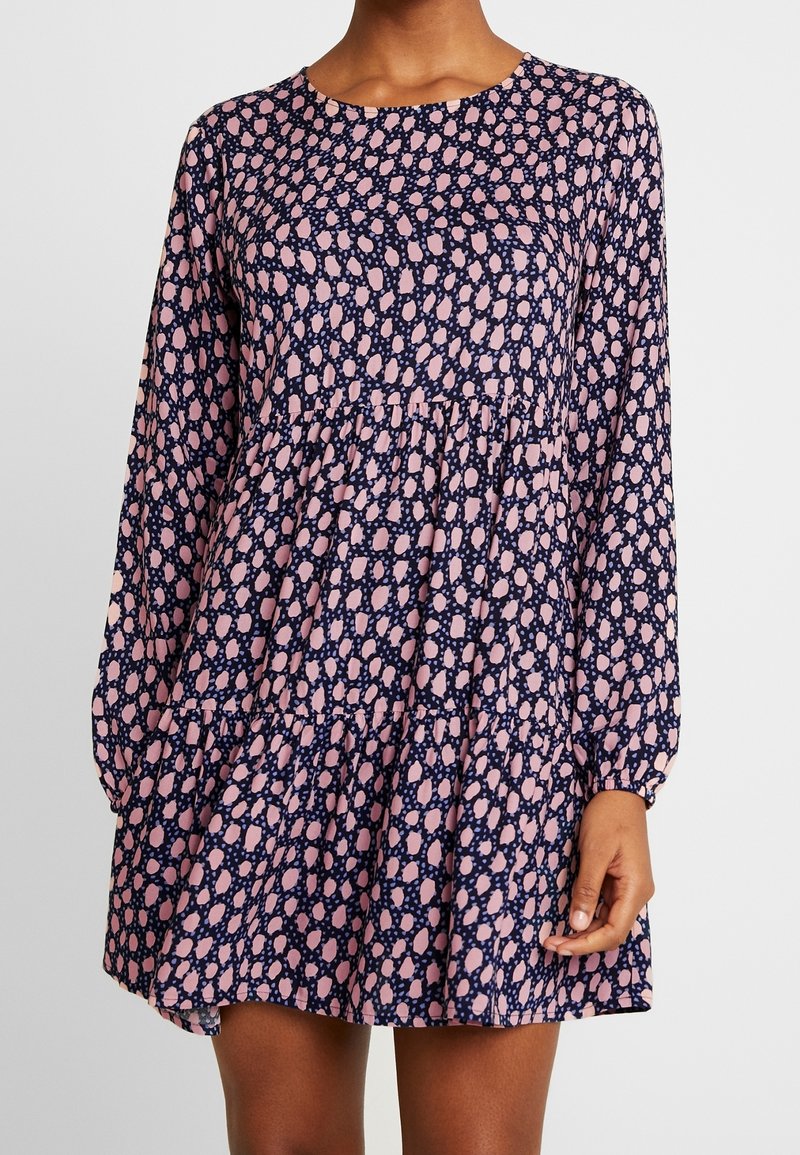 Mörkblå klänning med långa ärmar, rund halsringning och en volangkant i flera lager. Har ett rosa abstrakt mönster genom hela. Slät tygtextur.