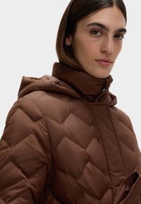 Brun quiltet pufferjakke med hætte, der har et chevronmønster, høj krave og lynlås foran. Glat tekstur.