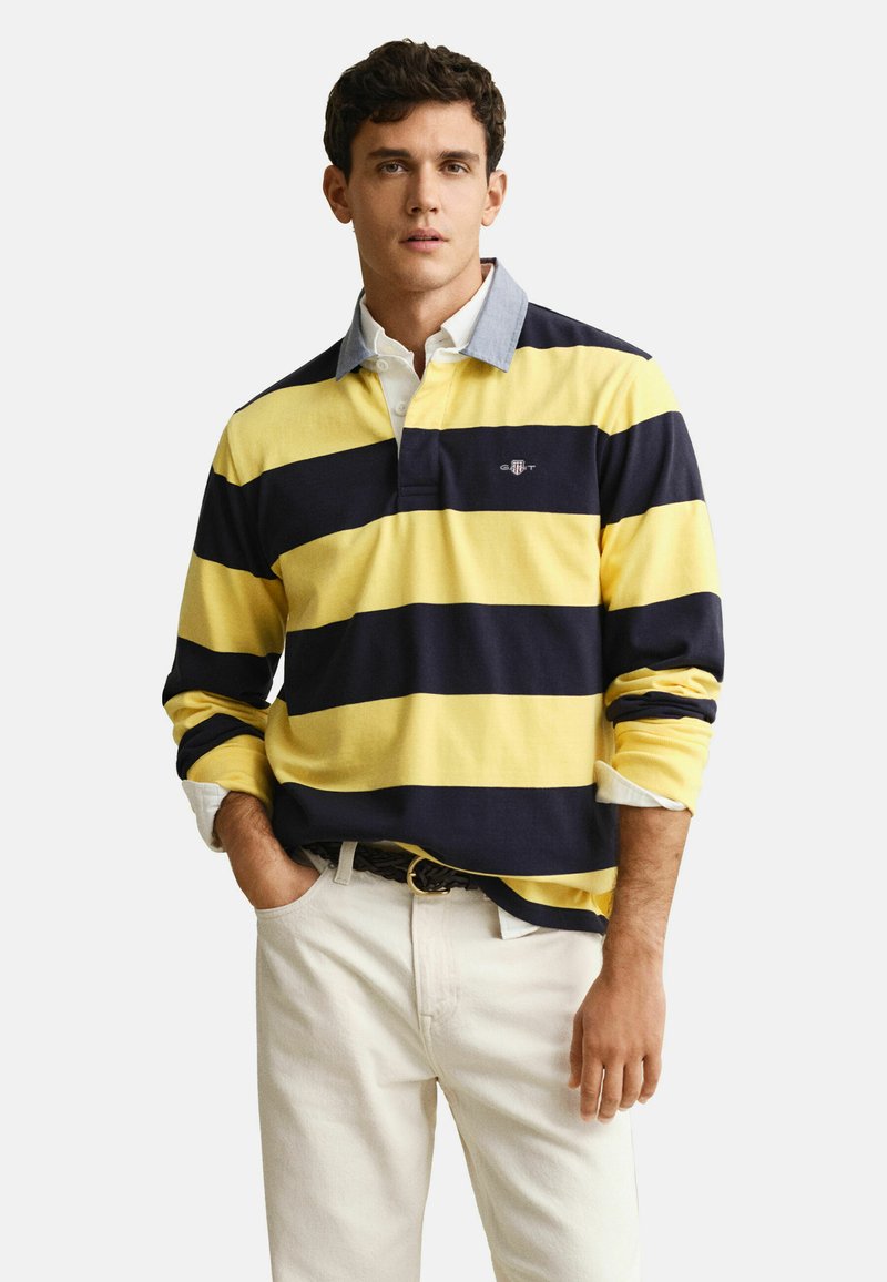 Mann trägt ein gelb-navy gestreiftes Rugbyshirt über einem weißen Hemd mit Kragen, dazu helle Hosen, steht mit einer Hand in der Tasche.