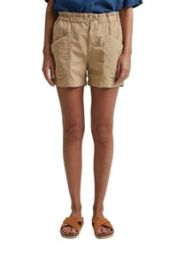 Femme portant un short beige à taille élastique avec poches avant et des sandales marron à brides croisées, debout les mains le long du corps.