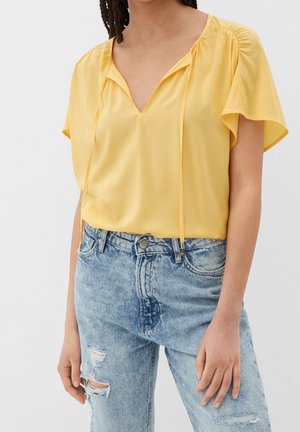 Kobieta ubrana w luźną żółtą bluzkę z krótkim rękawem i dekoltem w serek oraz jasnoniebieskie podarte dżinsy z wysokim stanem.