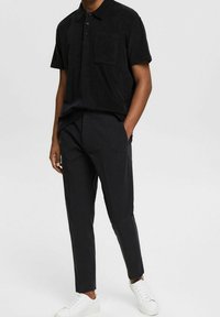 Homme portant un polo noir à manches courtes, un pantalon noir ajusté avec les mains dans les poches, et des baskets blanches, se tenant devant un fond uni.