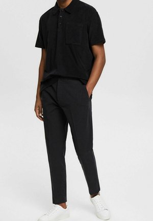 Mann trägt ein schwarzes Polohemd mit kurzen Ärmeln, schwarze eng anliegende Hosen, hat die Hände in den Taschen und weiße Sneakers, steht vor einem einfarbigen Hintergrund.