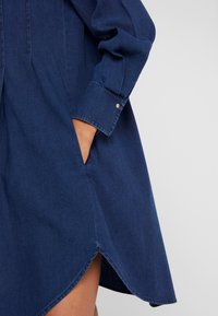 Džínové šaty v tmavě modré barvě s dlouhými rukávy, boční kapsou a dlouhou spodní částí, která se mírně zakřivuje.