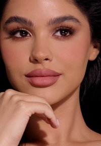 Primer plano de un rostro con piel suave y clara, labios mate en tono malva, cejas definidas y cabello oscuro y ondulado, con una mano descansando sobre la barbilla, fondo neutro.