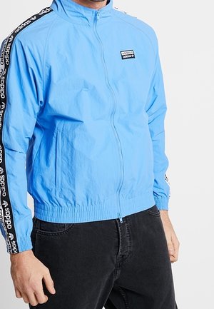 Homme portant une veste de sport Adidas bleu clair avec des bandes noires du logo sur les manches et un pantalon noir, debout les mains détendues.