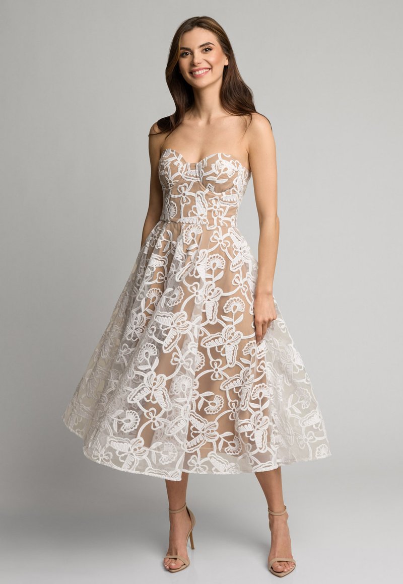 Bezramionowa biała sukienka z kwiatowym haftem, dopasowanym topem i rozkloszowanym dołem. Przezroczysta narzutka z misternymi wzorami z koronką i wyraźnie zaznaczoną talią.