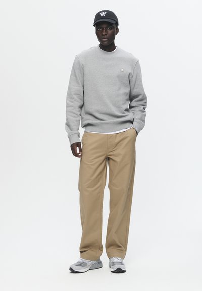 Felpa grigia con polsini a coste, pantaloni beige e sneakers grigie. Il cappellino è nero con un logo in lettere bianche. Outfit semplice e casual.