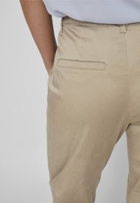 Pantaloni beige strutturati con una texture liscia e una tasca posteriore singola. Presentano una vita media e un dettaglio di cucitura sottile.