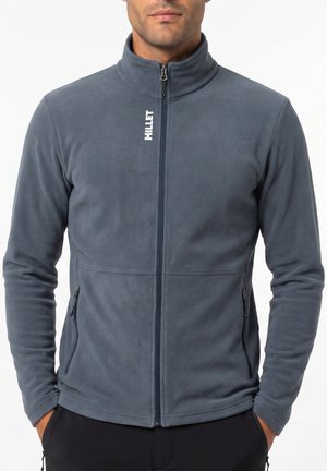 Man wearing a gray zip-up fleece jacket with two zippered side pockets and the word "MILLET" printed vertically near the collar.