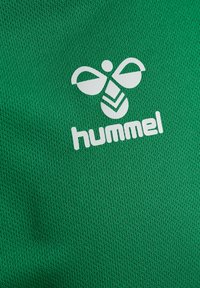 Zielony materiał z delikatnym wzorem teksturowym, z wyraźnym białym logo oraz abstrakcyjnym wzorem przypominającym pszczołę nad nazwą marki "hummel."