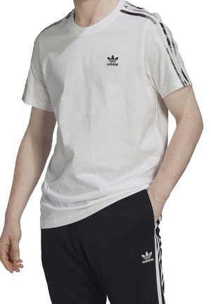 Valkoinen puuvillapaita, jossa on musta Adidas-logo rinnassa ja mustat vaakasuorat raidat olkapäillä, yhdistetty mustiin housuihin.