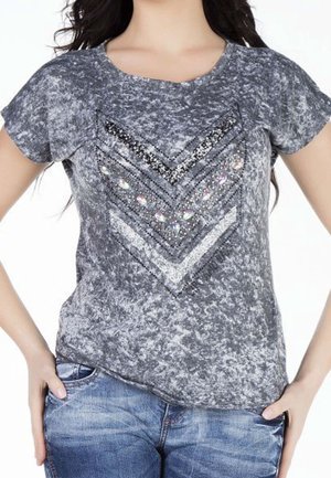 Frau trägt ein kurzärmeliges grau marmoriertes Shirt mit verzierten Chevron-Mustern und blaue Jeans, Hände ruhen in den Taschen.