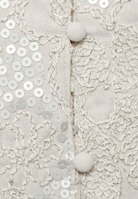 Svetlý textil s detailným kvetinovým výšivom. Obsahuje kruhové strieborné flitre, textúrovanú priadzu a detail gombíka pokrytého tkaninou.