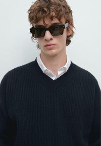Maglione nero a coste con scollo a V, indossato sopra una camicia bianca con colletto. Occhiali da sole neri a forma rettangolare con lenti trasparenti.
