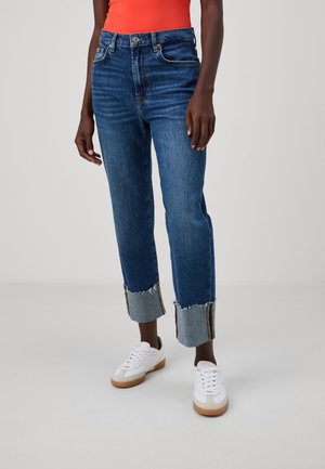 Osoba ubrana w ciemnoniebieskie jeansy z mankietami oraz postrzępionymi krawędziami, białe trampki i pomarańczową bluzkę, stojąca na tle jednolitego, jasnego tła.