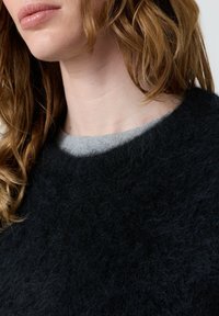 Moteris su šviesia oda ir banguotais šviesiai rudu plauku, vilkinti pūkuotą juodą megztinį ant pilkos marškinės, kirptos žemiau smakro ir pečių.