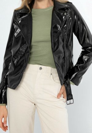 Kvinde iført skinnende sort læder bikerjakke over grøn ribbet skjorte og højttaljede off-white bukser, hånd i lommen.
