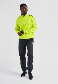Pullover giallo fluorescente con zip e accenti neri sulle spalle, abbinato a pantaloni jogger neri con logo e polsini elastici. Scarpe nere.