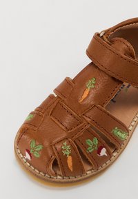 Sandale en cuir marron avec orteils découverts et plusieurs sangles. Présente de vibrantes broderies végétales, y compris des carottes et des radis, sur le dessus.