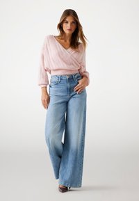 Gaiši rozā adīts džemperis ar V-veida izgriezumu un balona formas piedurknēm, kombinēts ar augsta vidukļa plaša piegriezuma zilām džinsa biksēm. Tīrs dizains, ikdienišķa piegriezuma.