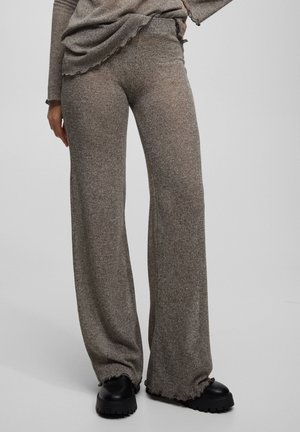 Femme portant un pantalon marron ample en maille côtelée, associé à des chaussures noires épaisses, debout devant un fond clair uni.