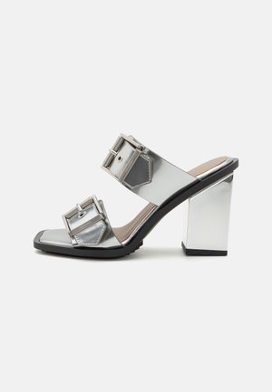 Metallic silver high-heeled mules with wide straps and large rectangular buckles, featuring a square toe and chunky mirrored heel.