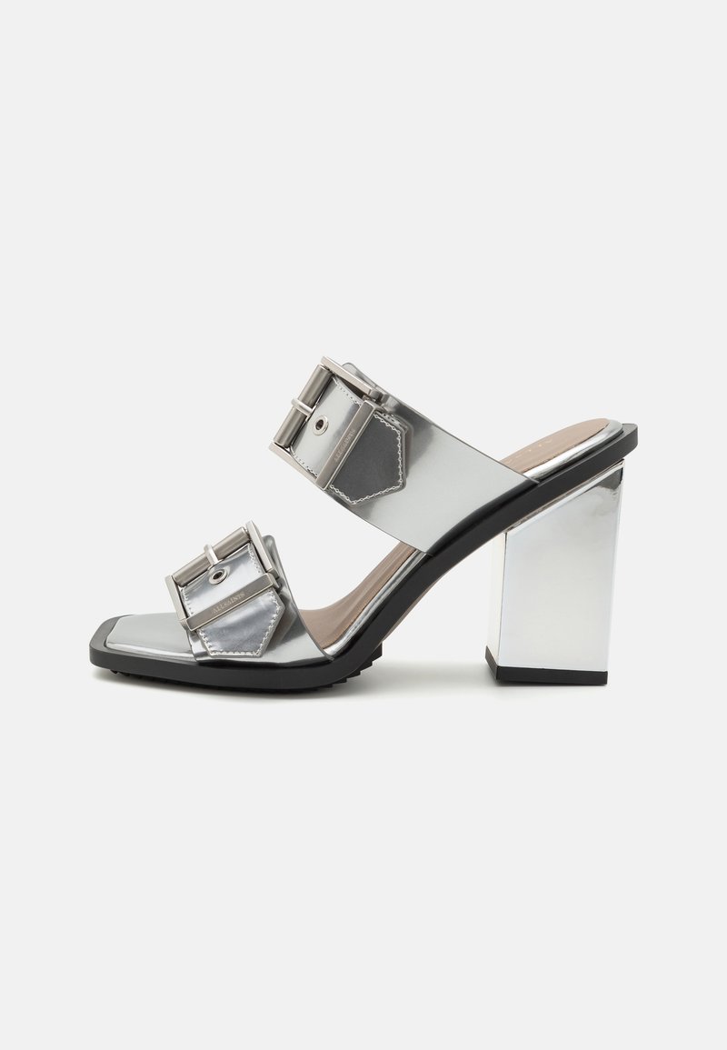Mules à talons hauts argentés métalliques avec larges bracelets et grandes boucles rectangulaires, présentant un bout carré et un talon épais miroir.