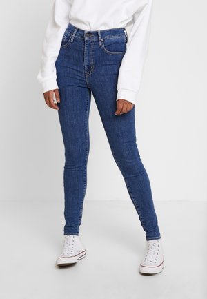 Pantalones vaqueros ajustados de mezclilla azul con una cintura alta, con trabillas para el cinturón, bolsillos frontales y un ligero estiramiento. Llevados con zapatillas altas blancas.