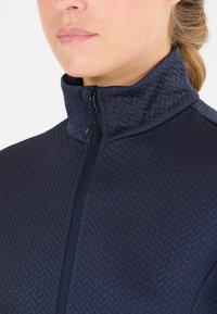 Marinblå zip-jacka med ett strukturerat vävt mönster, hög krage och dragkedjedetaljer, tillverkad av ett mjukt, elastiskt material.