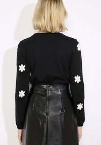 Jersey negro con apliques de flores blancas en las mangas, combinado con una falda de cuero negro brillante, que resalta una silueta elegante.