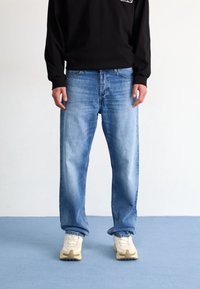 Jeans de denim azul claro con un corte relajado, que presentan un desvanecimiento sutil. Combinados con una camiseta negra de manga larga y zapatillas color crema.