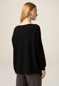 Czarny, dziergany sweter o luźnym kroju, z teksturowanym wzorem, szerokim dekoltem i długimi rękawami. Noszony z szarymi spodniami.