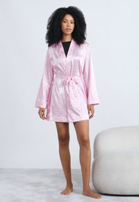 Κοντό ρόμπα από ροζ σατέν με μακριά μανίκια, ζώνη στη μέση και λεία υφή. Το ύφασμα διαθέτει διακοσμητικά σχέδια.