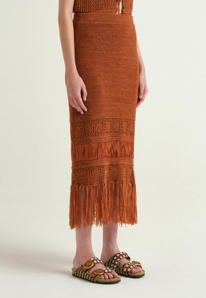 Donna che indossa una gonna midi in maglia color ruggine con dettagli a frange e sandali slip-on con perline e grandi fibbie circolari.