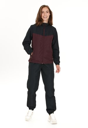 Windbreaker-Jacke in Schwarz und Maroon, mit hohem Kragen, Frontreißverschluss und Reißverschlusstaschen. Kombiniert mit schwarzen Jogginghosen.