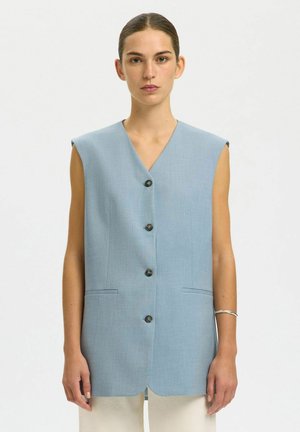Frau trägt ärmellose hellblaue, mit Knöpfen versehene Weste mit V-Ausschnitt und zwei aufgesetzten Taschen, steht mit neutralem Gesichtsausdruck vor weißem Hintergrund.