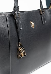 Czarna torebka o teksturowanej powierzchni z gładkim wykończeniem, wyposażona w złote okucia i zawieszkę z metalową plakietką z logo. Dwa górne uchwyty do noszenia.