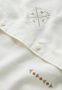 Valge särk, millel on keerukad tikitud mustrid: geomeetriline disain helehallis ja peened kolmnurkse mustriga täpid halli ja oranži toonides.