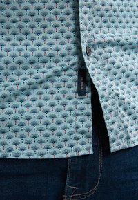 Camicia di cotone azzurro chiaro con un motivo di forme a ventaglio e accenti rossi. Presenta una chiusura con bottoni e un'etichetta sulla cucitura laterale.