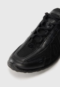 Črn športni copat z mrežasto in usnjeno površino, zavezanim sprednjim delom in teksturirano gumijasto podplato, prikazan od blizu na svetlem ozadju.