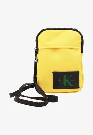 Bolso bandolera amarillo de Calvin Klein Jeans con cremallera negra, parche con logo negro y correa negra ajustable.