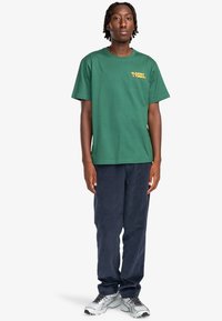 Green cotton t-shirt with short sleeves and yellow graphic text on the chest, paired with navy corduroy pants and silver athletic shoes.