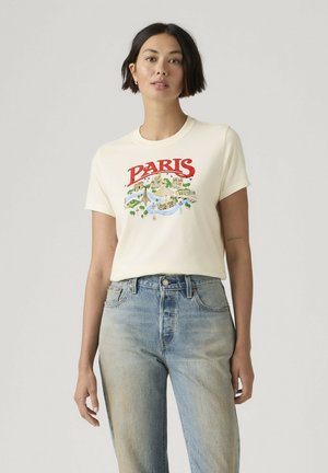 Femme aux cheveux courts et foncés portant un t-shirt blanc avec un imprimé rouge de Paris et un jean bleu clair sur un fond uni clair.