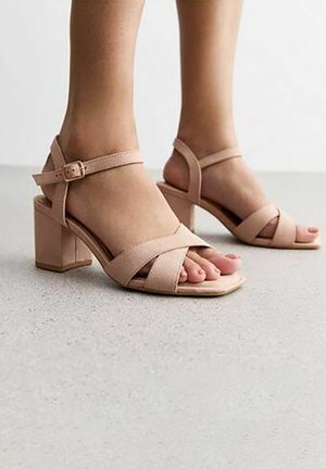 Pieds portant des sandales à talons blocs ouvertes aux orteils, de couleur rose blush, avec des brides à la cheville, sur un sol gris clair.