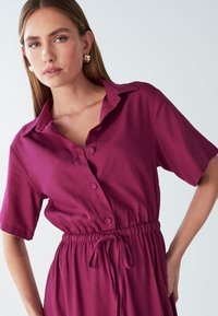 Žena nosi kratku haljinu s kratkim rukavima, dugmadi u boji šljive, s pojasom na vezanje i zlatnim okruglim naušnicama, na jednostavnoj svijetloj pozadini.