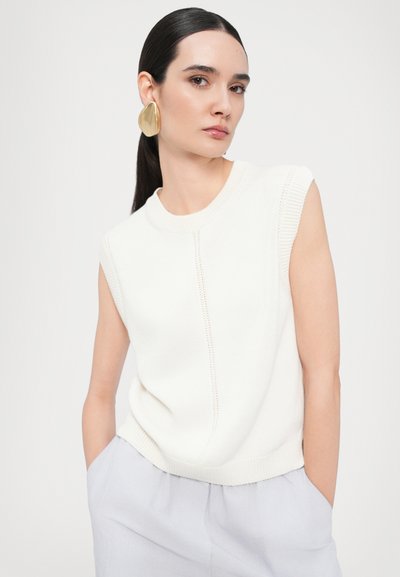 Maglione bianco senza maniche in maglia con scollatura a costine, caratterizzato da dettagli di cuciture verticali. La modella indossa orecchini d'oro grandi e astratti e pantaloni chiari.