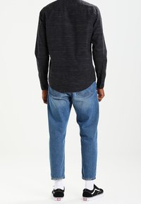 Uomo che indossa una maglietta a maniche lunghe nera, jeans blu, calzini bianchi e sneakers nere, in piedi girato di spalle su uno sfondo bianco.