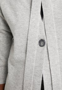 Cardigan gris clair en tissu tricoté doux, avec une fermeture à boutons sur le devant, un design ouvert et des manches trois-quarts.