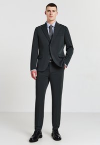 Completo scuro su misura realizzato in tessuto liscio, con giacca a un petto e rever a taglio, abbinato a una camicia azzurro chiaro e una cravatta con motivo.